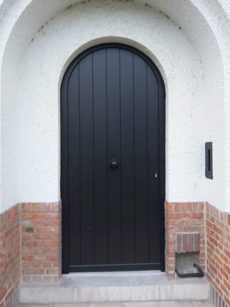 Porte d'entrée cintrée noire en bois à lames verticales avec bouton de tirage rond, sous une arche crépie blanche